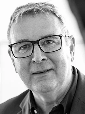 Schwarz-Weiß-Foto eines Mannes mit Brille, kurze Haare, lächelnd in einem dunklen Anzug