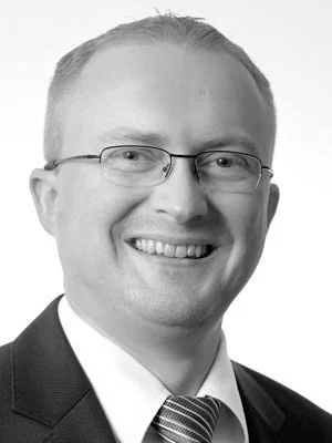 Schwarz-weiß Porträt eines Mannes mit Brille, Anzug und Krawatte, lächelnd.