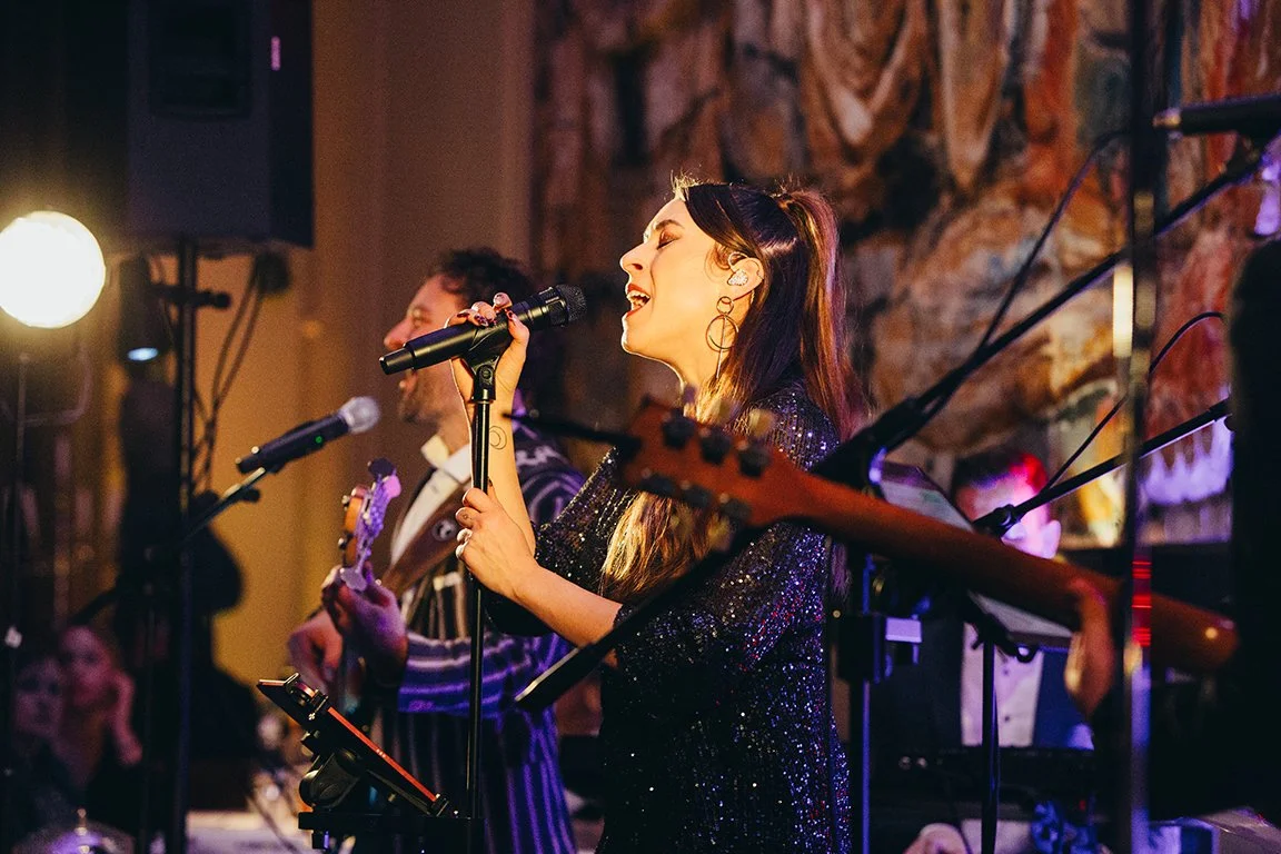 Eine Frau singt leidenschaftlich mit einem Mikrofon, im Hintergrund spielt eine andere Person Gitarre, bei einer Live-Musik-Performance in einem gemütlichen Raum mit Holzdekoration.