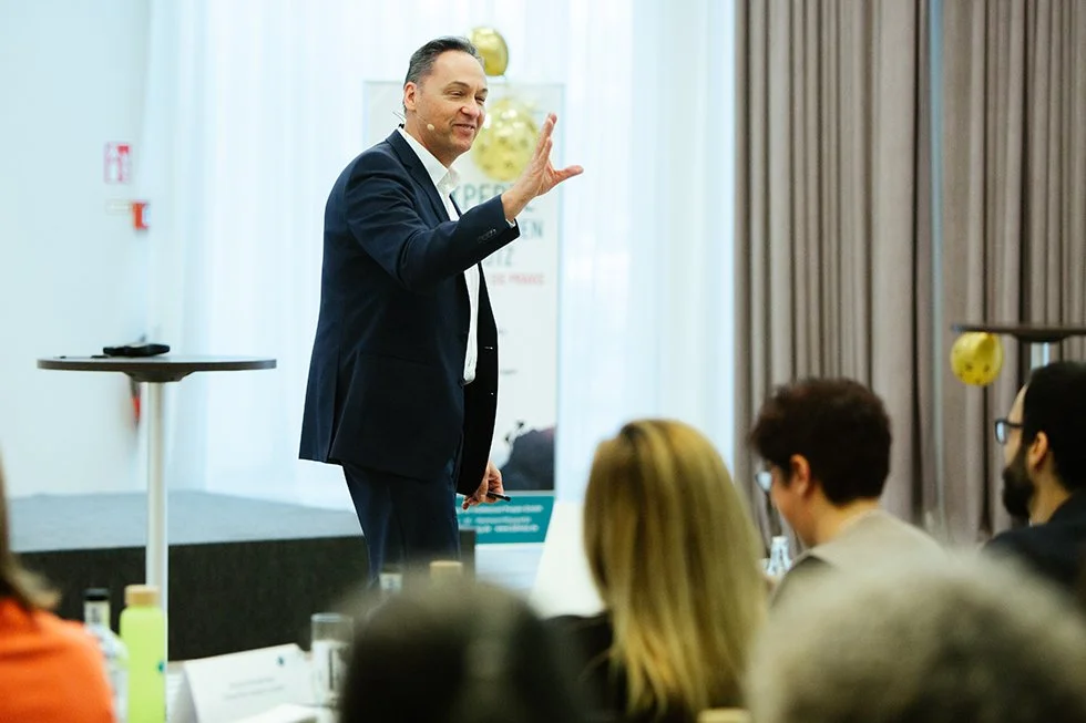 Ein Mann in einem dunklen Anzug spricht vor einem Publikum in einem Konferenzraum.
