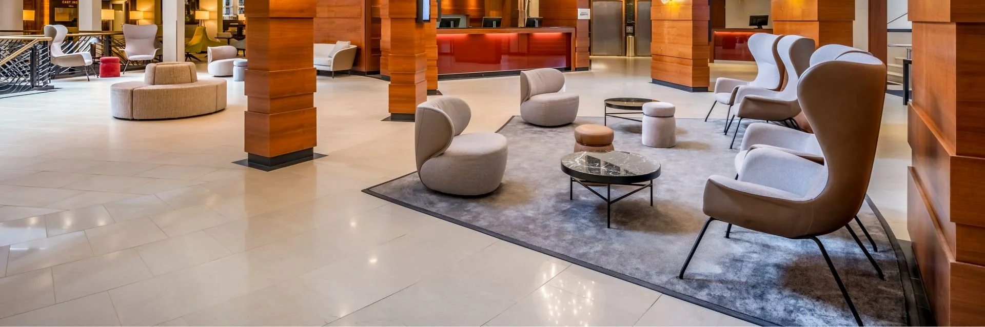 Innenaufnahme einer Hotellobby mit bequemen Stühlen und Tischen auf einem grauen Teppich, Holzverkleidungen und eine Rezeption im Hintergrund.