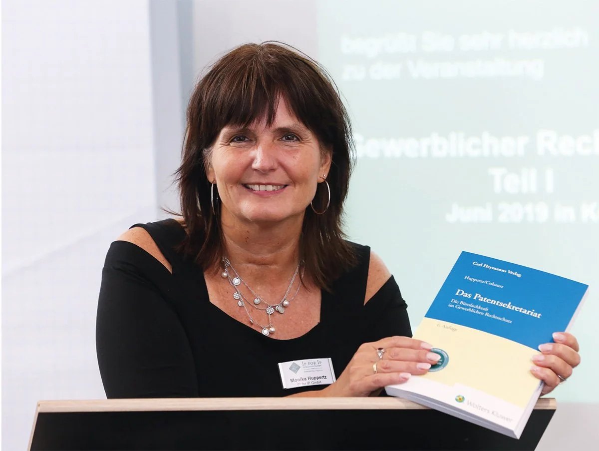 Eine Frau hält ein Buch mit dem Titel 'Das Patentsekretariat' in einer Vorlesung oder Präsentation.