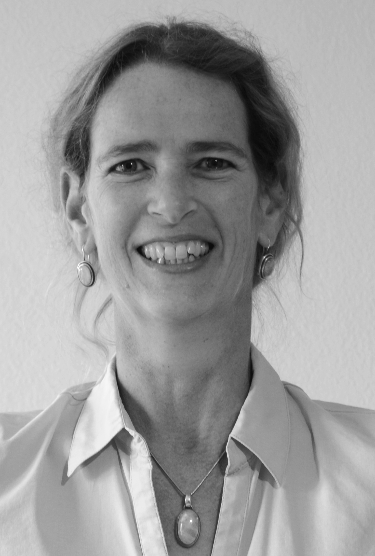 Schwarz-weiß-Foto einer lachenden Frau, die eine weiße Bluse und auffälligen Schmuck trägt, vor einer neutralen Wand.