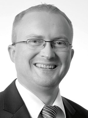 Schwarz-weiß Porträt eines Mannes mit Brille, Anzug und Krawatte, lächelnd.