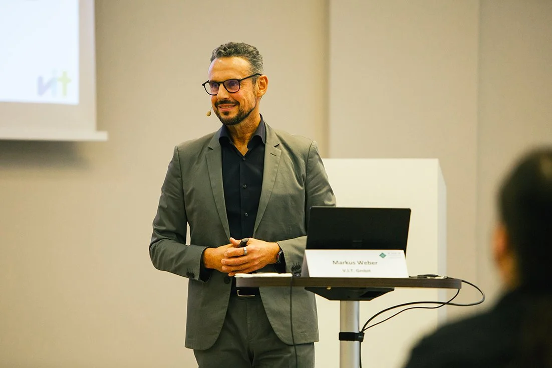 Mann in grauem Anzug bei einer Präsentation, steht vor einem Laptop und einem Projektor, lächelt, trägt eine Brille.