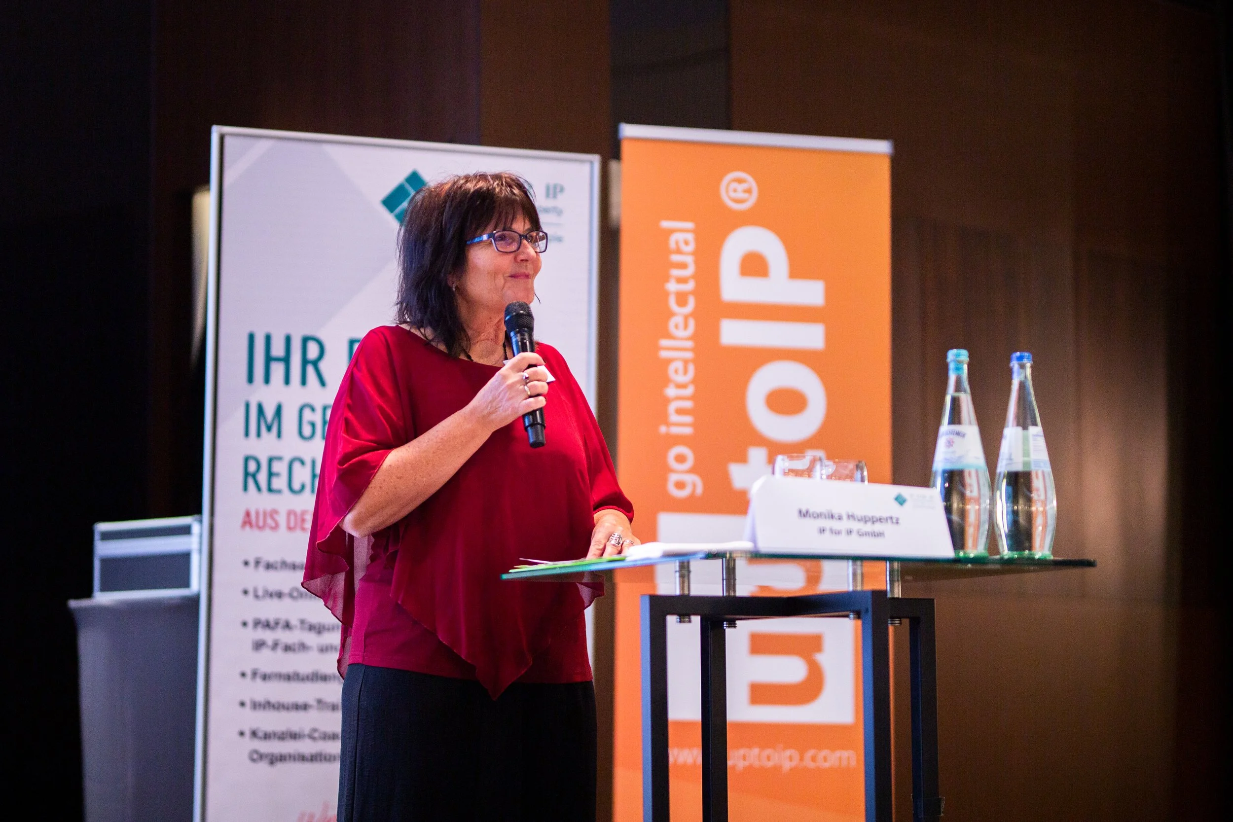 Eine Frau mit Brille und roten Oberteil spricht bei einer Veranstaltung. Hinter ihr sind Banner und Tische mit Wasserflaschen sichtbar.