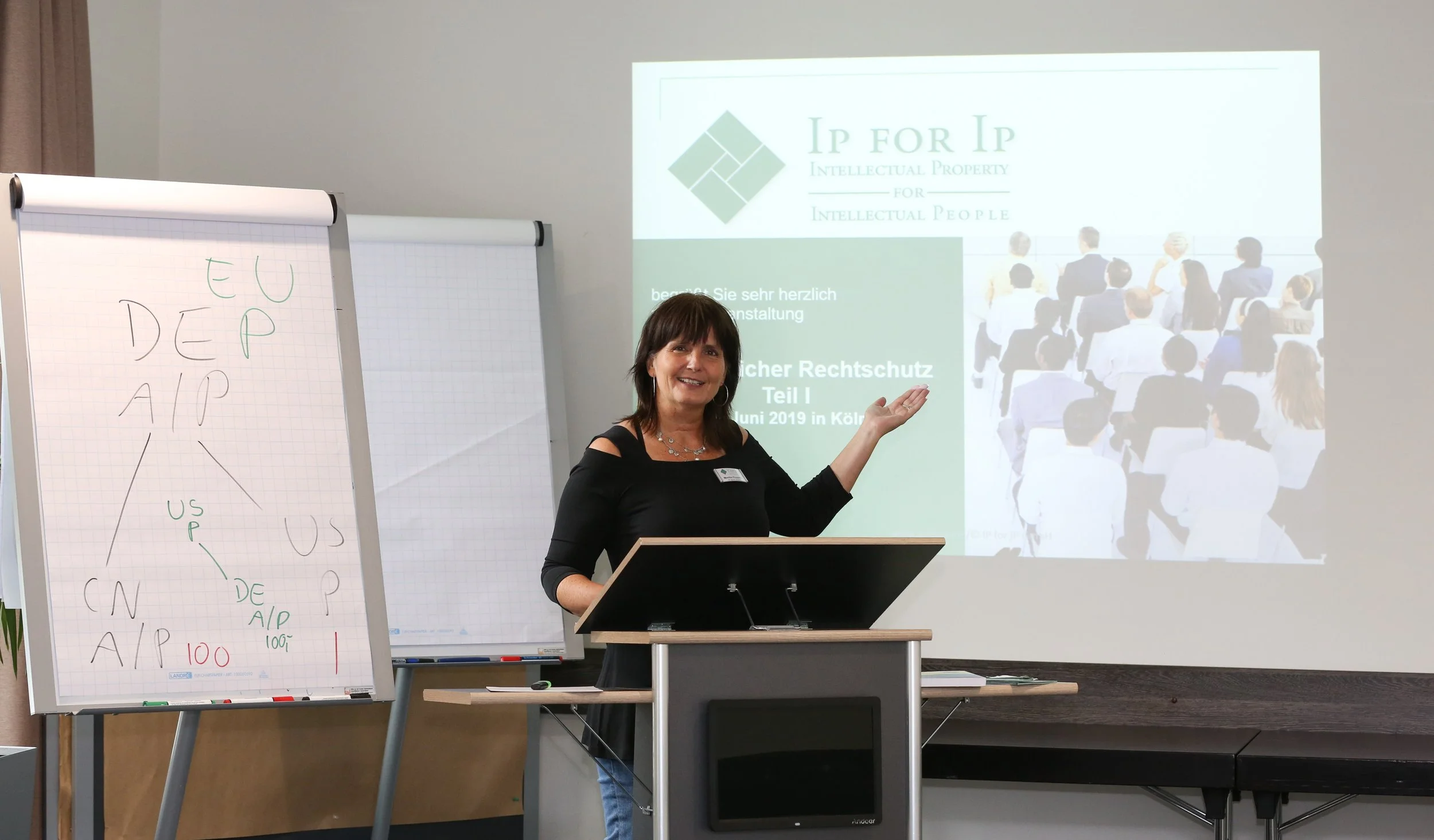 Eine Frau steht an einem Pult vor einer Präsentation, während im Hintergrund auf einer Leinwand ein Logo und Text zum Thema geistiges Eigentum angezeigt werden. Neben ihr steht eine Flipchart mit Diagrammen und Text.