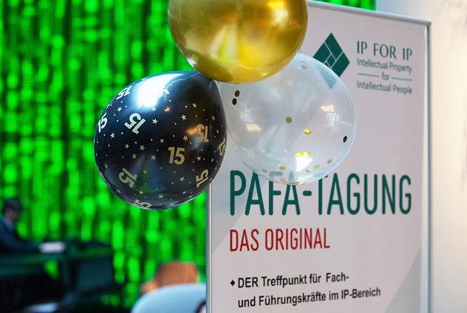 Blaue, weiße und goldene Ballons vor einem Poster der IP For IP Veranstaltung.