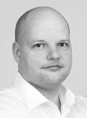 Schwarz-weiß-Porträt eines Mannes mit kurzer, glatter Kopfhaut, trug ein weißes Hemd, schaut ernst in die Kamera.