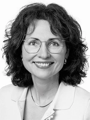 Eine lächelnde Frau mit curly Haaren und Brille, trägt eine weiße Jacke.
