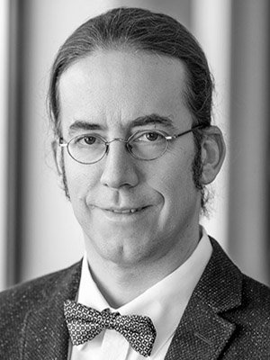 Schwarz-Weiß-Porträt eines Mannes mit Brille, Anzug und Fliege, lächelnd, vor einem neutralen Hintergrund.