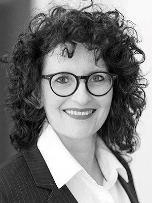 Schwarz-weiß-Foto einer Frau mit lockigem Haar, Brille und Business-Kleidung, die lächelnd in die Kamera schaut.