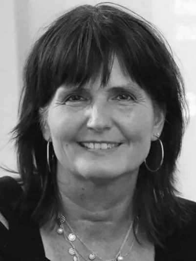 Schwarz-weiß-Foto einer lächelnden Frau mit dunklem, schulterlangem Haar, großen Ohrringen und Halsketten.