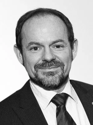 Schwarz-weiß-Fotografie eines Mannes in formeller Kleidung mit Anzug und Krawatte, lächelnd.