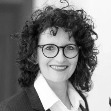 Porträt einer Frau mit lockigen Haaren und Brille, die lächelnd in die Kamera schaut.