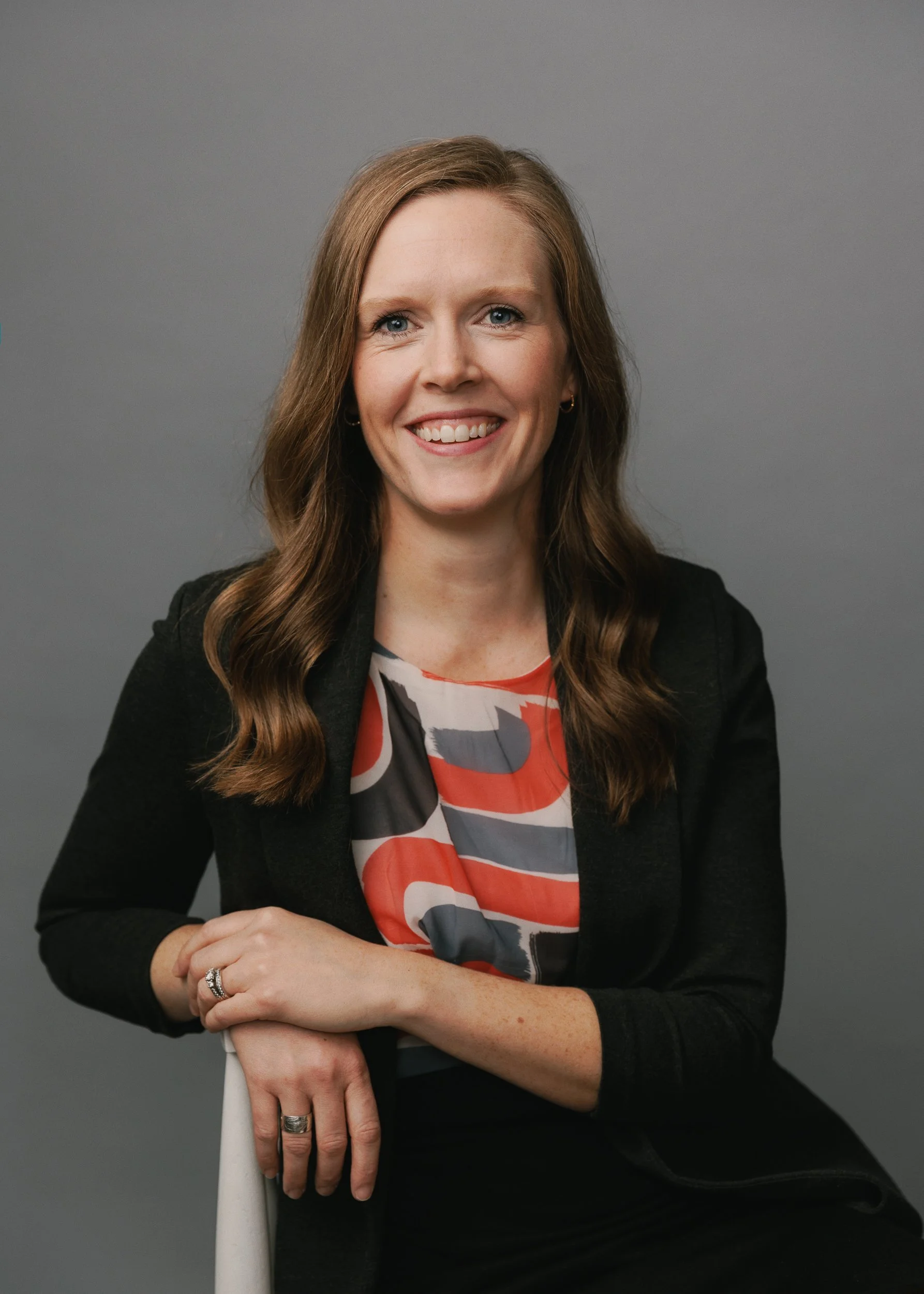 A woman with long wavy brown hair, wearing a black blazer and a colorful patterned blouse, sitting with her arms crossed, smiling against a gray background.