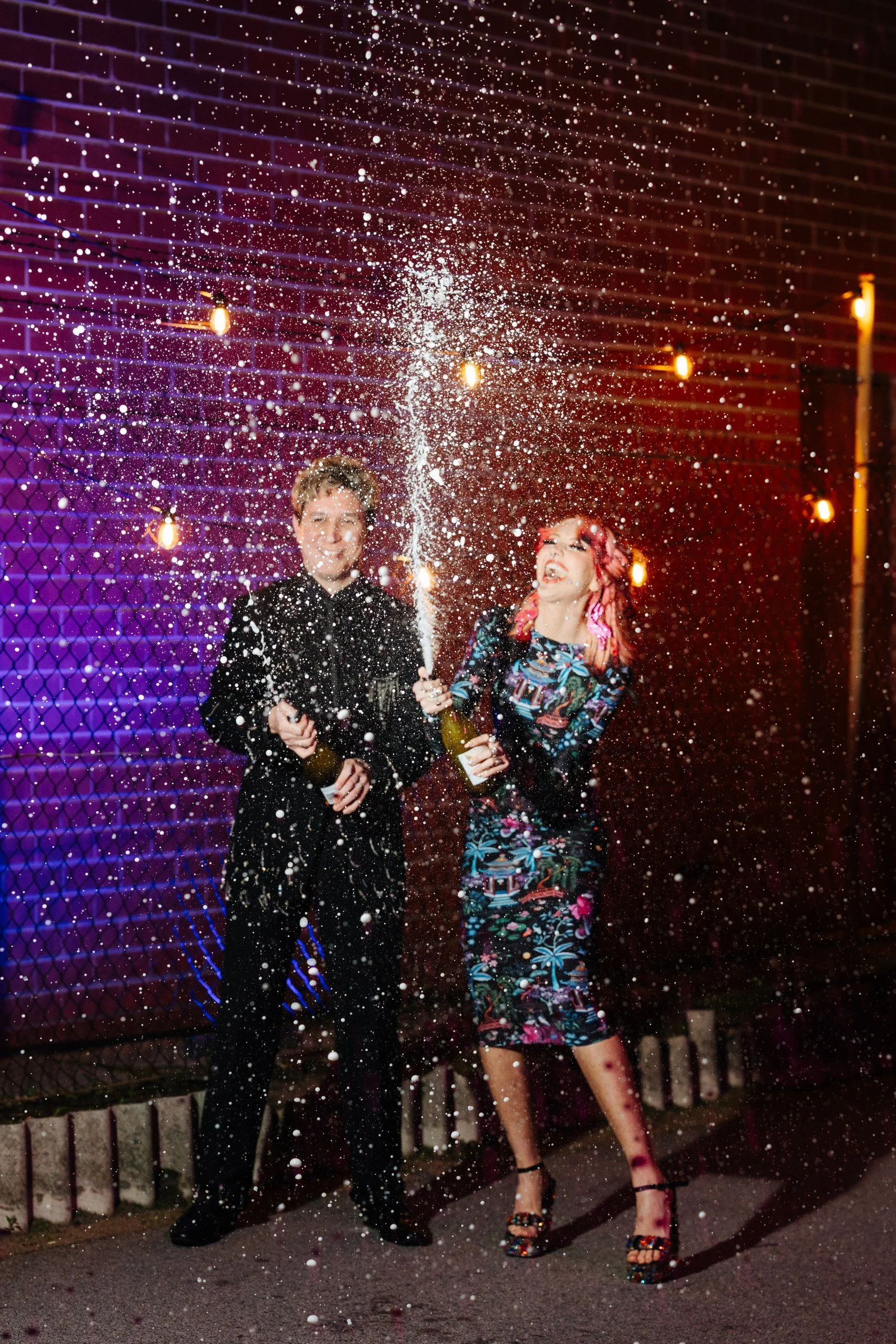 A man and woman celebrating with champagne in front of a colorful lit background at night. They are spraying champagne, with champagne droplets in the air.