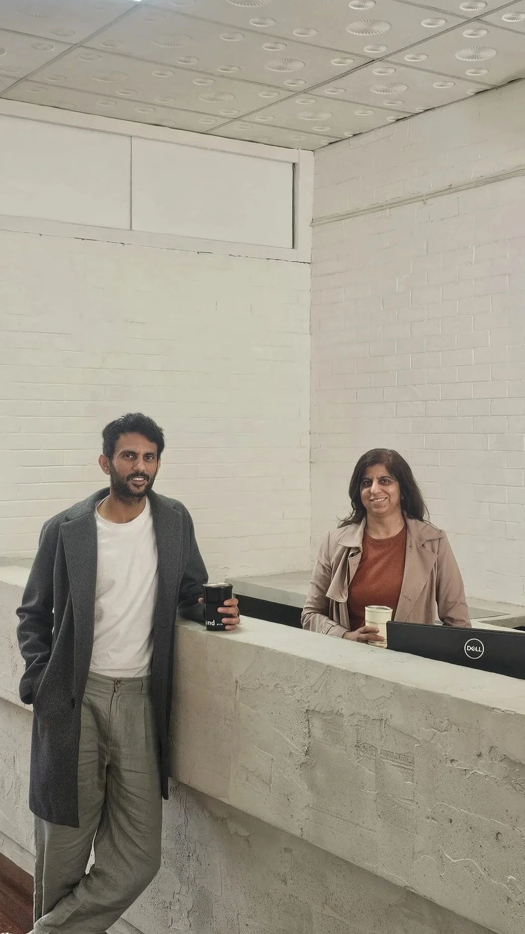 A man and woman standing at a reception desk, holding drinks, in a minimalistic room with white brick walls and a tiled ceiling.