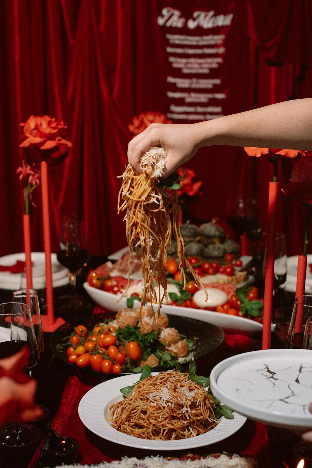 A hand holding spaghetti above a table with various dishes, including cherry tomatoes, meatballs, and salads, with red candles and a red curtain backdrop.