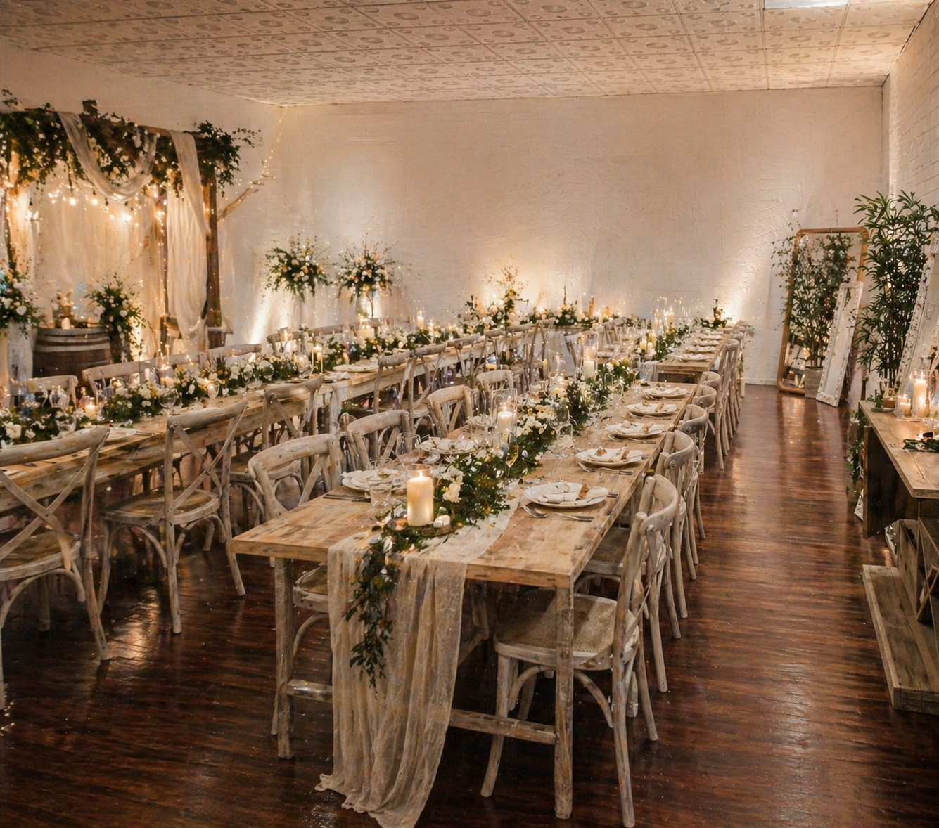 A decorated wedding reception hall with rustic wooden tables and chairs, floral arrangements, candles, and string lights, with a backdrop of draped fabric and greenery.