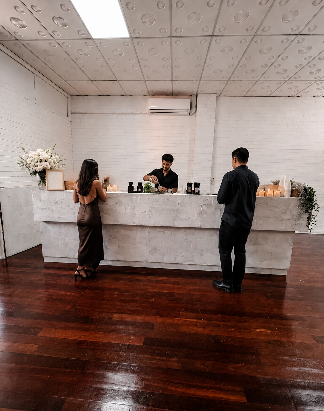 A woman and a man are at a bar counter, with a bartender preparing drinks. The woman is wearing a satin brown dress, and the man is dressed in black. The bar area has candles, flowers, and drink bottles, with a white brick wall and a wooden floor in the background.