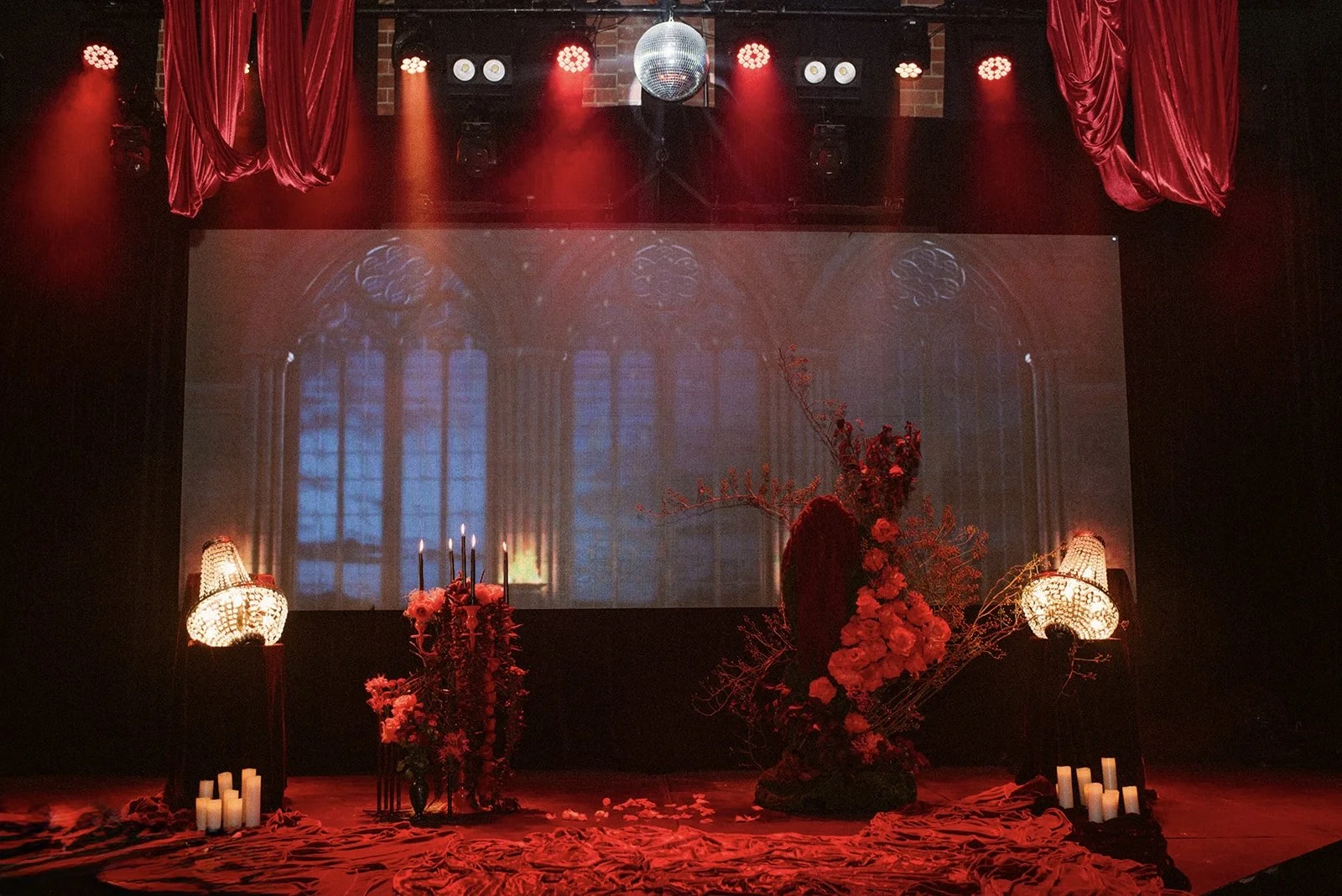 Stage decorated with red flowers, candles, and chandeliers, with a backdrop of a Gothic window projection and red curtains overhead.