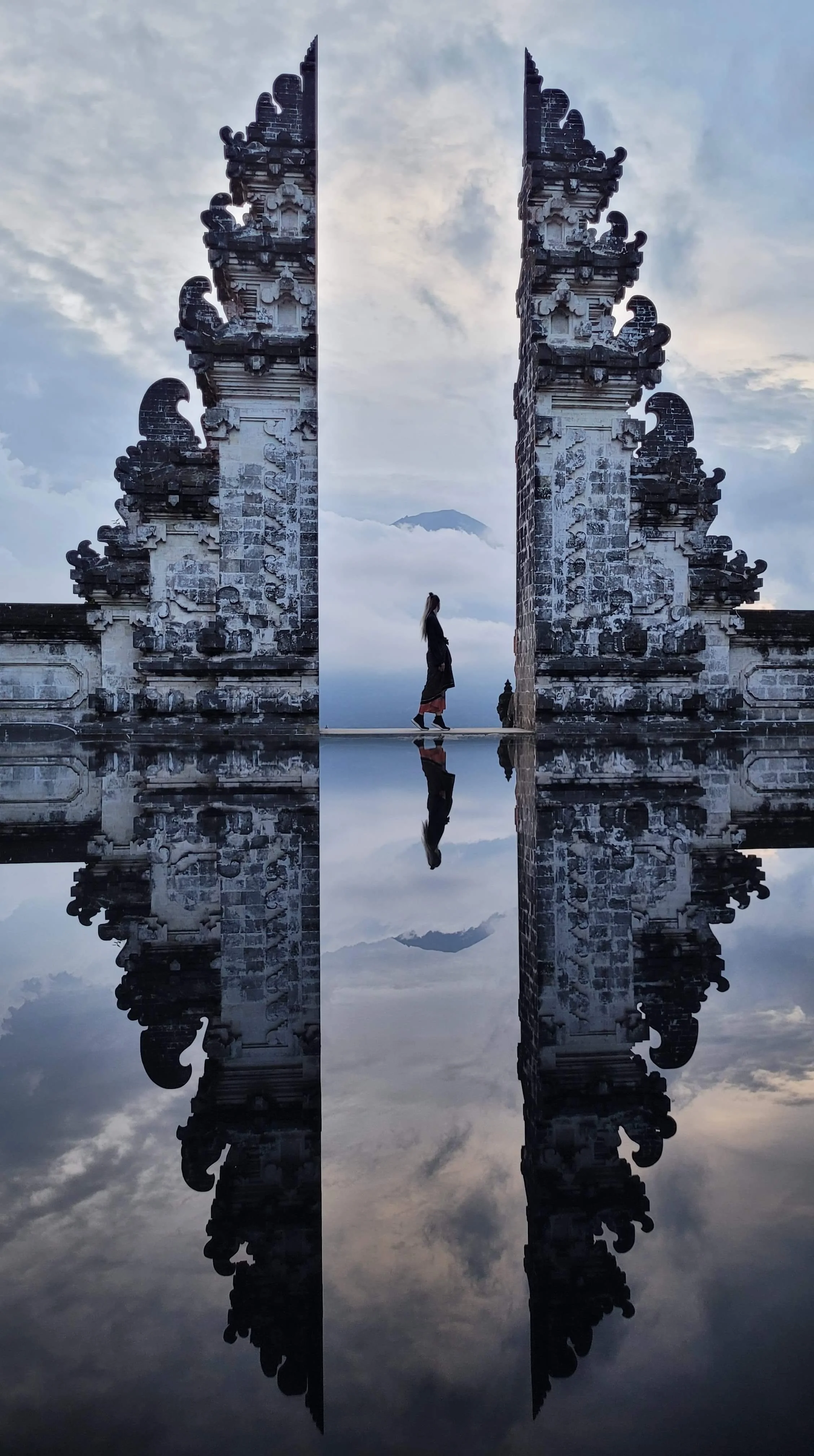 A person walking through an intricately carved stone gate with a mountain and cloudy sky in the background, reflected in a body of water below.