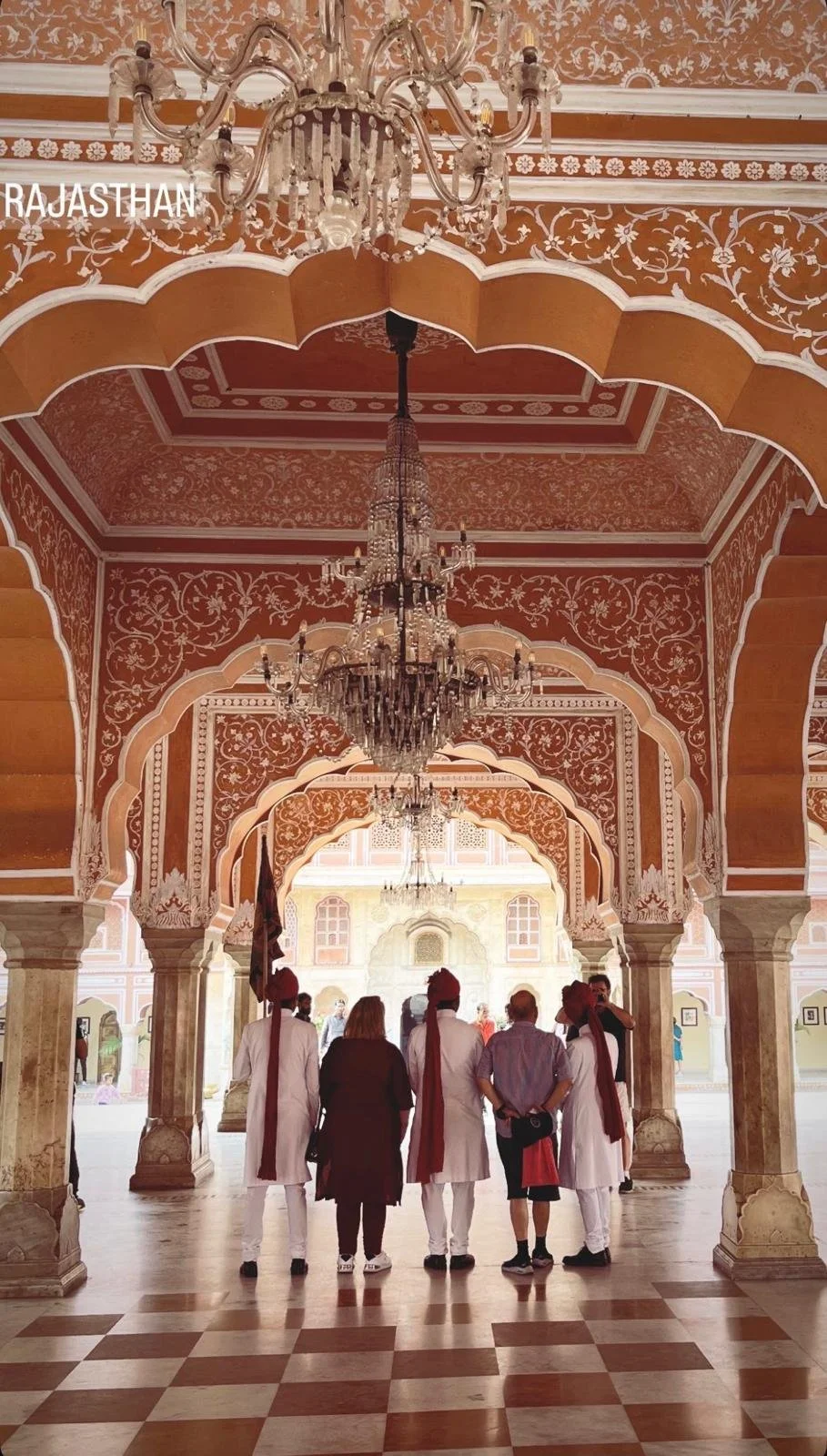 People walking under and looking at grand chandeliers in a decorated palace corridor with ornate arches and intricate wall designs, in Rajasthan.
