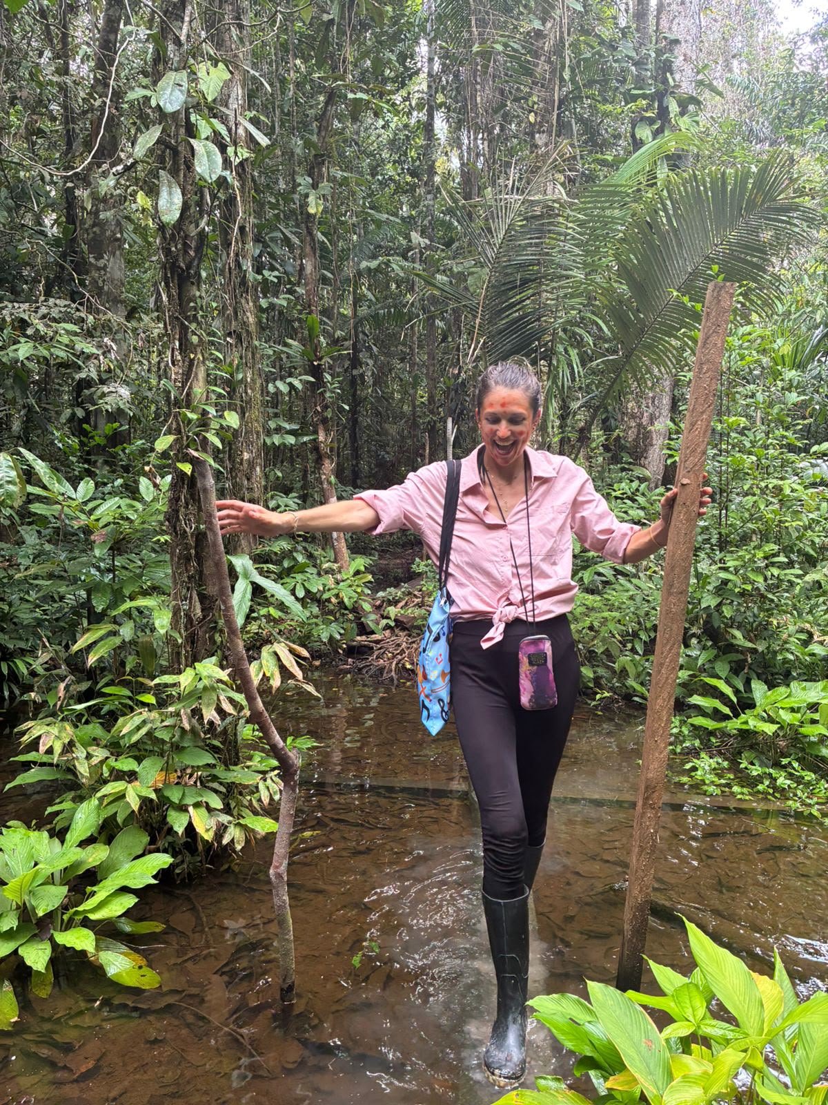 A woman in a pink shirt and black pants walking through a shallow stream in a lush green jungle, holding a wooden stick for support and smiling.