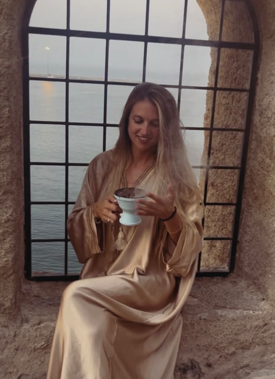 A woman with long blonde hair, wearing a shiny beige satin dress, is sitting on a ledge by a large window with a view of the water and sky. She is smiling and holding a small ceramic cup or bowl, looking at its contents.