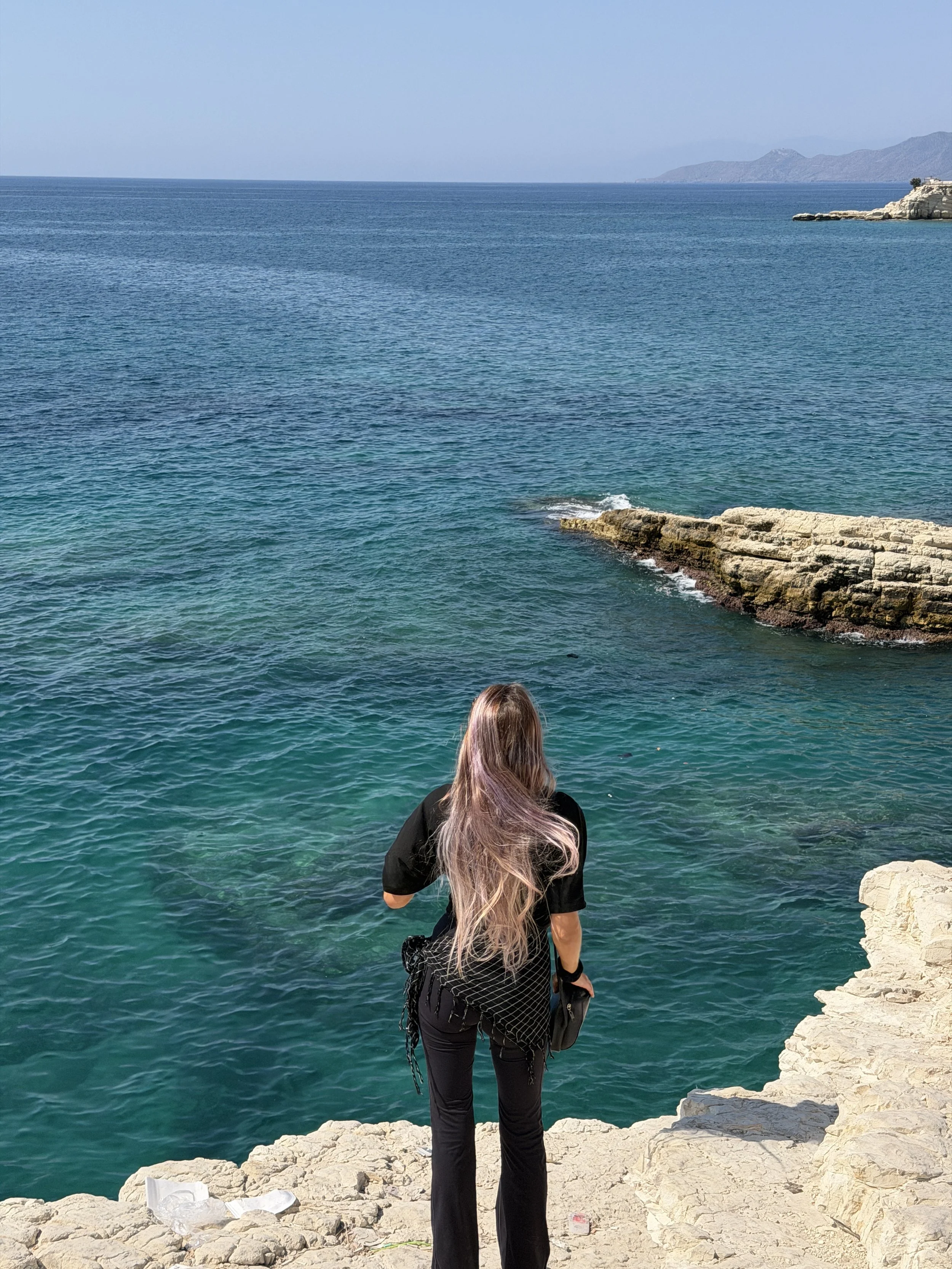 A person with long, light-colored hair standing on a rocky shoreline, facing the ocean with mountains in the background.