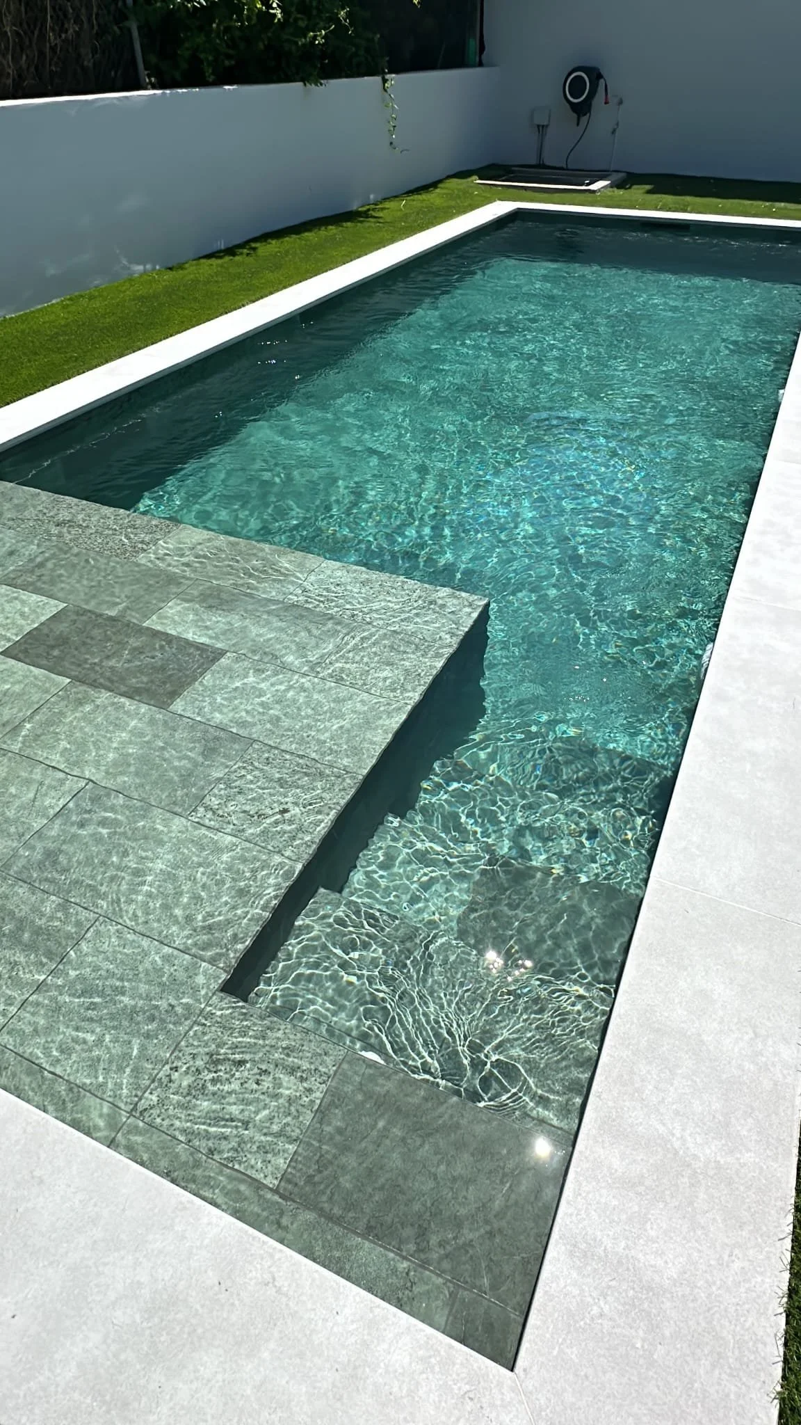 A modern outdoor swimming pool with clear blue water, gray stone tiles surrounding it, a grass strip along one side, and a white wall in the background with a black hose reel attached.