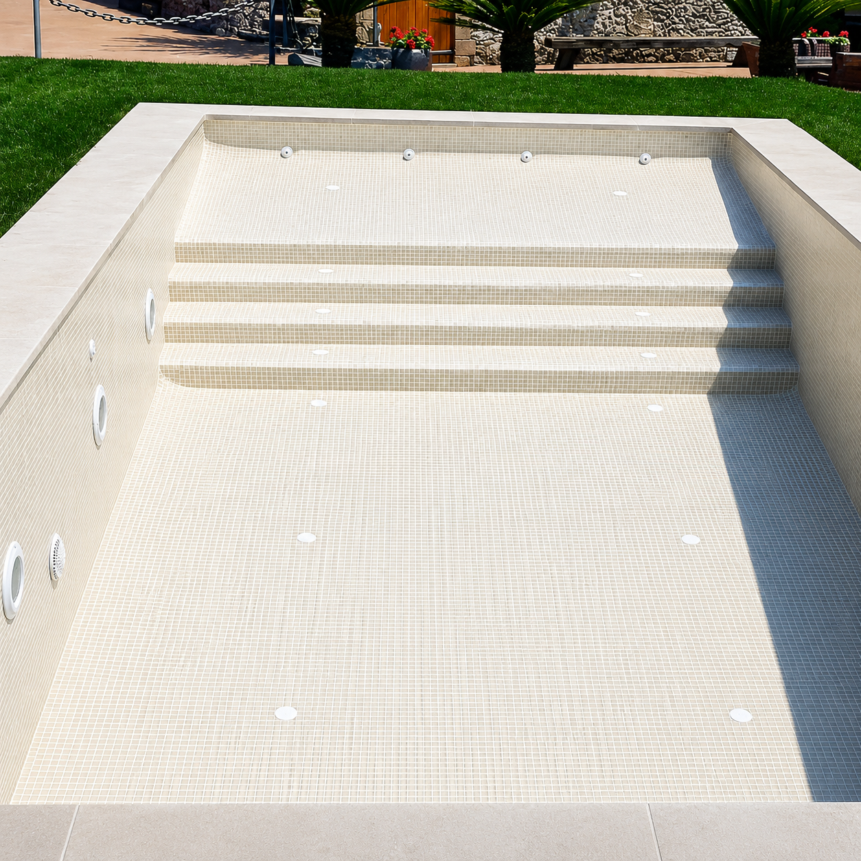 Empty swimming pool with beige tiles, stairs, and water jets, surrounded by green grass and outdoor furniture.