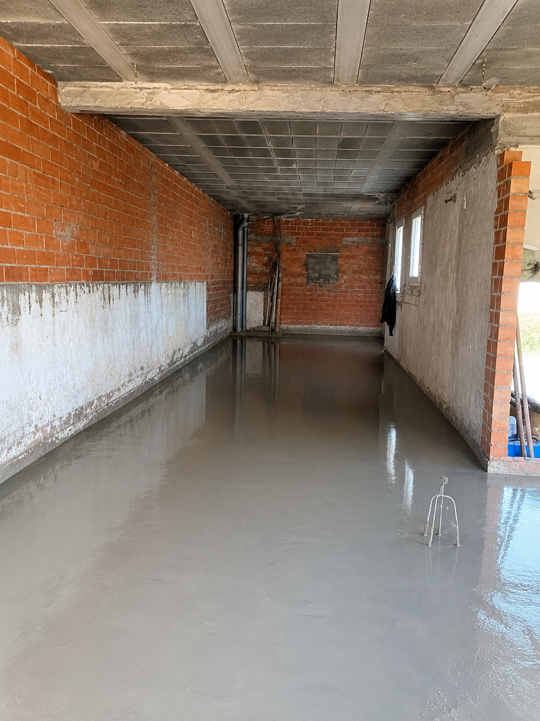 Interior of a building under construction with a freshly poured concrete floor, exposed brick walls, and a concrete ceiling. There are two small windows on the right wall and construction tools in the background.