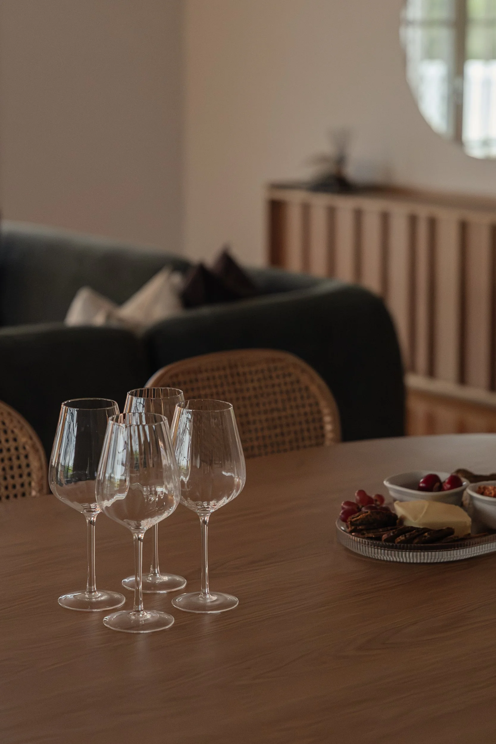 Empty wine glasses on a wooden table, with a plate of assorted snacks, including grapes, crackers, and cheese, in the background.