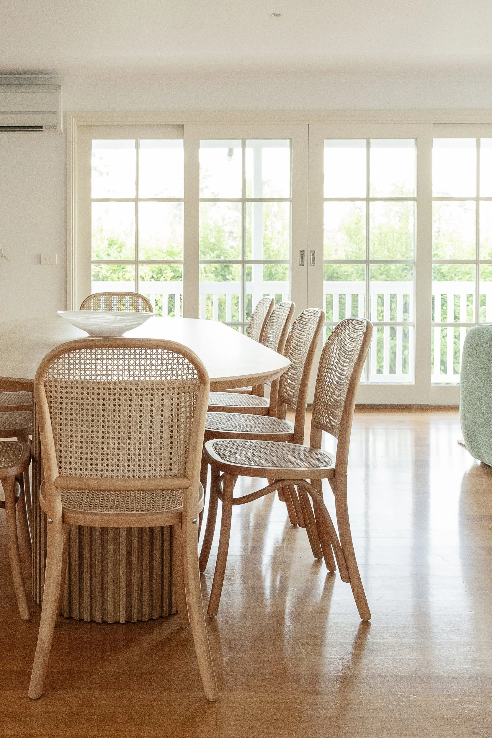 Dining room with a round wooden table surrounded by woven rattan chairs, a decorative ceramic bowl on the table, large glass-paneled sliding doors with white frames leading to a balcony with green foliage outside.