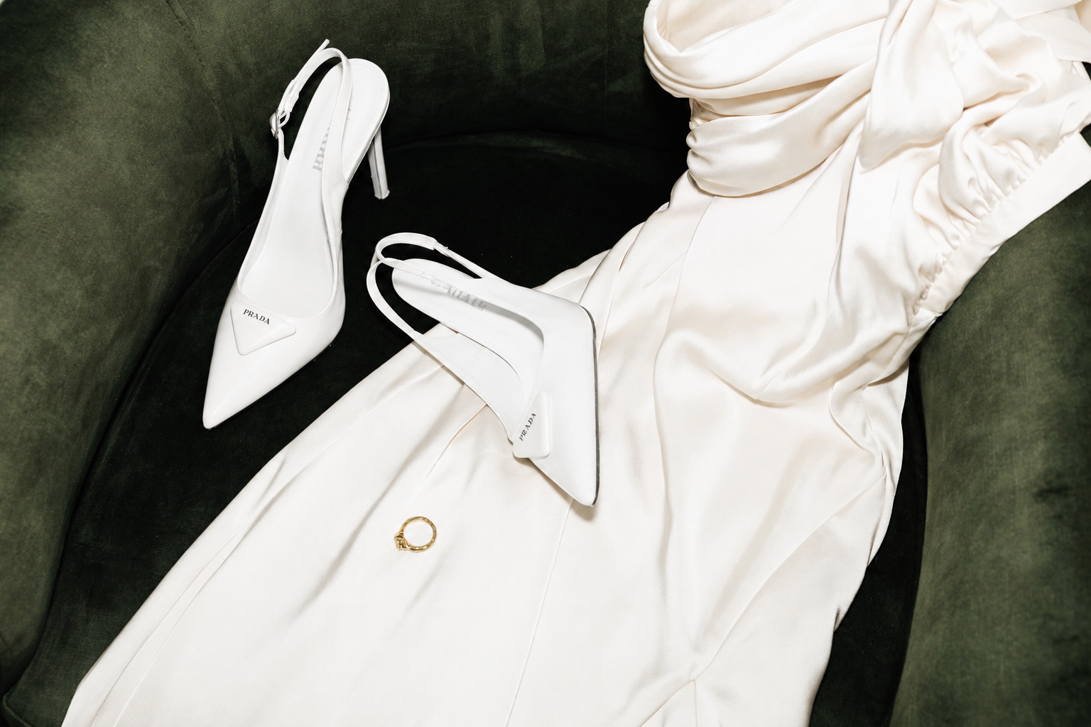 White Prada high heels, a gold ring, and a cream-colored satin dress or fabric on a green velvet chair.
