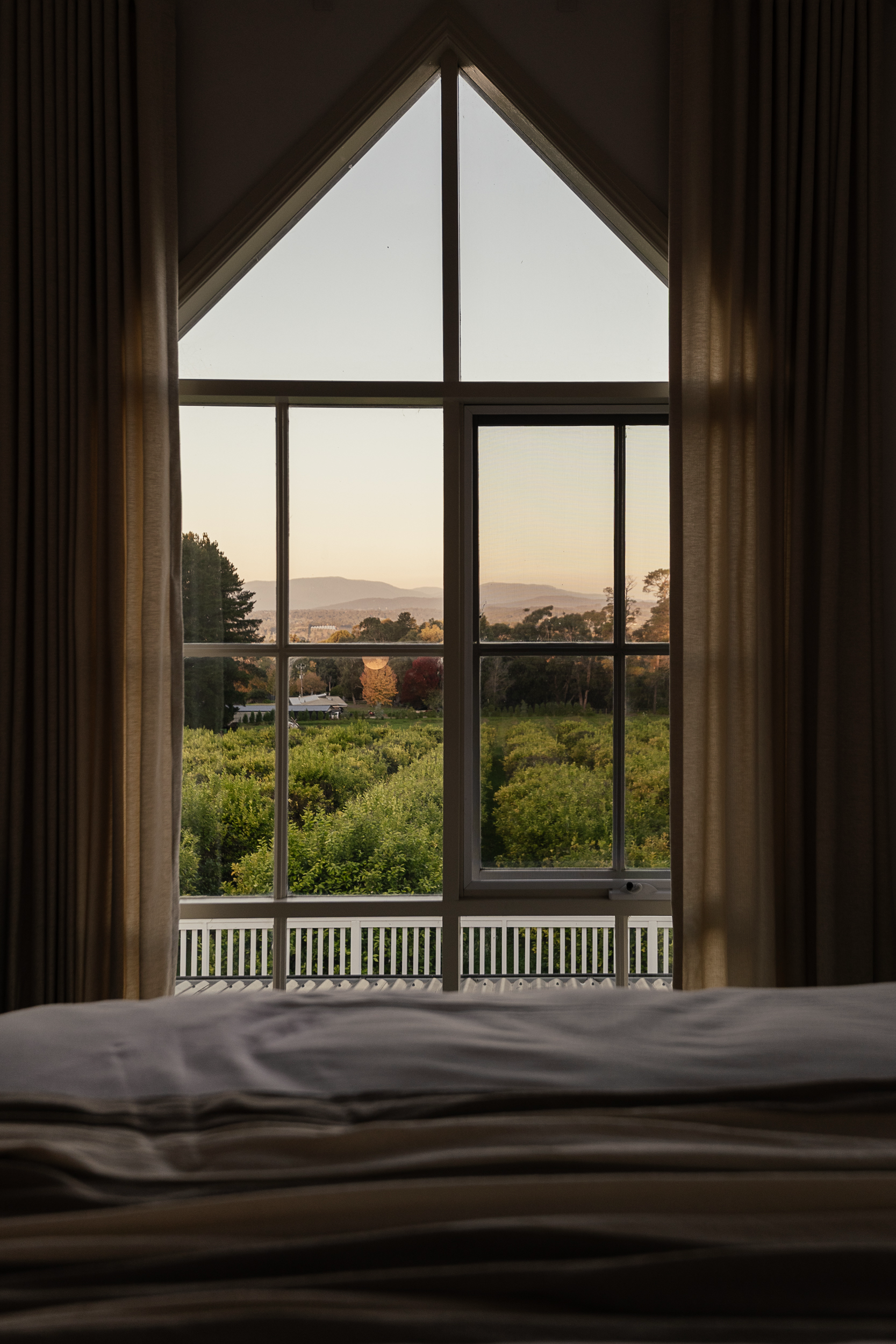 View from inside a room looking out a large window with curtains on either side. The landscape outside shows trees, a field, and distant mountains at sunset or sunrise.