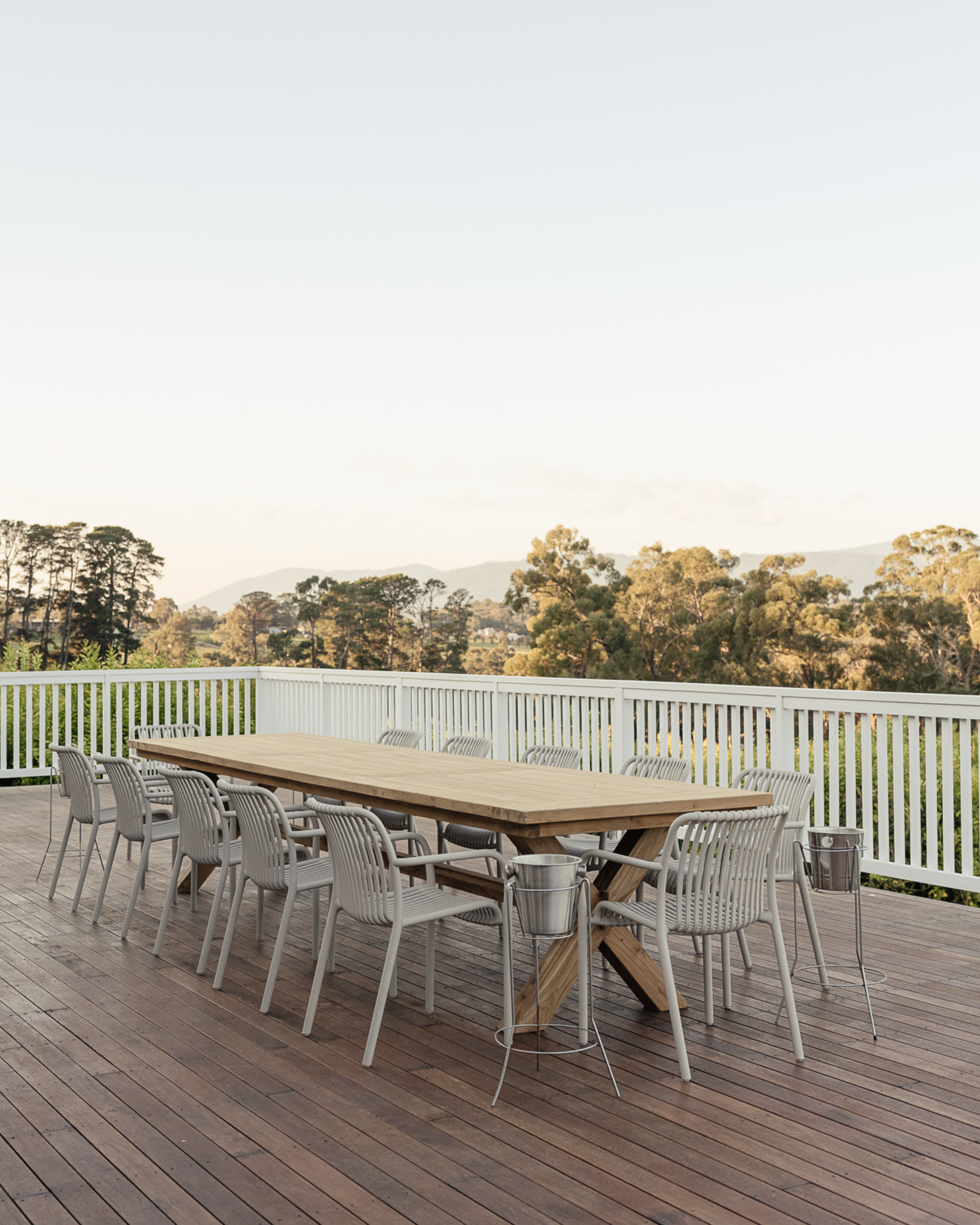 An outdoor wooden deck with a large rectangular wooden table surrounded by white chairs, two metal buckets on the ground, a white railing, and a scenic view of trees and distant mountains under a clear sky.