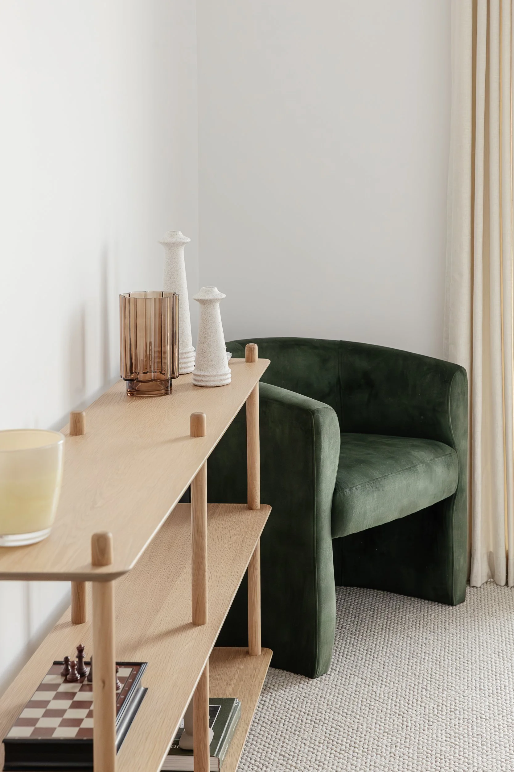 A light wooden shelf with decorative vases and a chessboard, positioned near a green velvet armchair in a room with white walls, cream curtains, and a textured beige carpet.