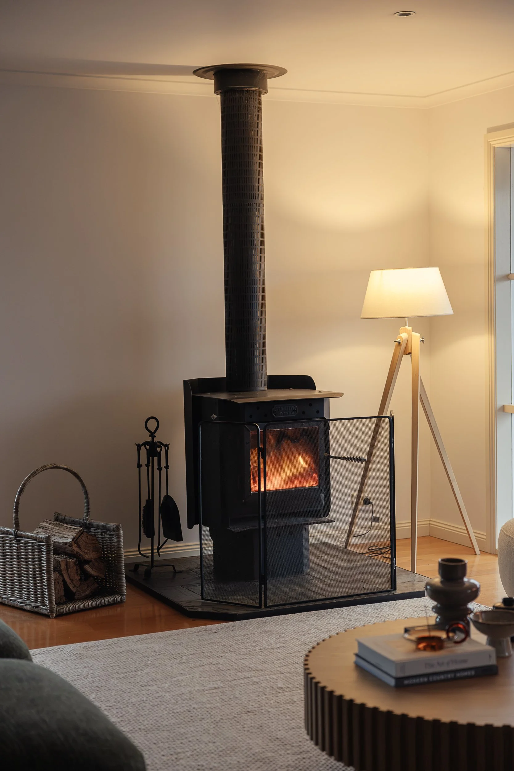 Living room with wood-burning stove, wicker basket of firewood, floor lamp, and a coffee table with books and decorative items, in a cozy and well-lit space.