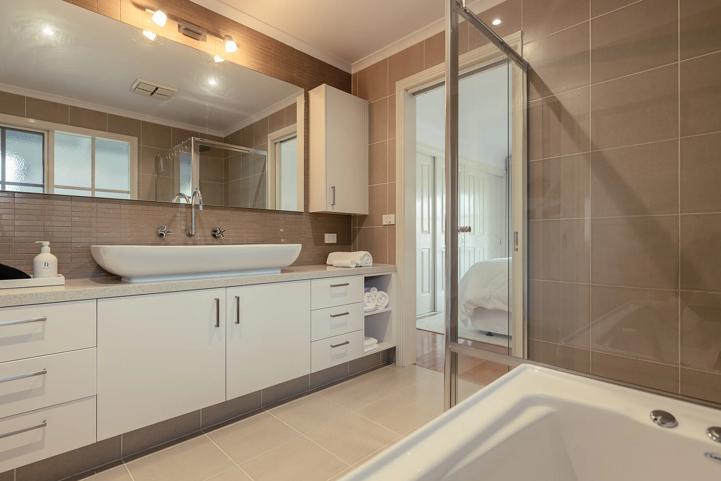 Bathroom with beige tiled walls, white vanity with sink, large mirror above, glass shower door, folded towels, and a window.