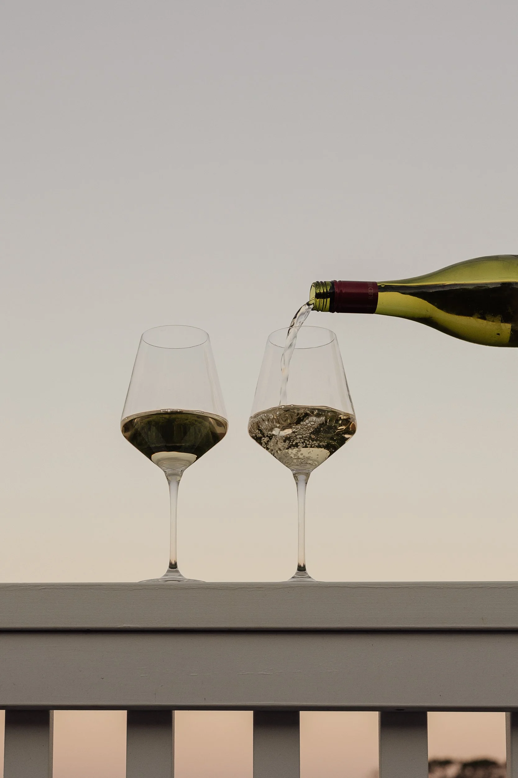 A bottle of white wine pouring into two wine glasses on a balcony railing against a soft gradient sky background.