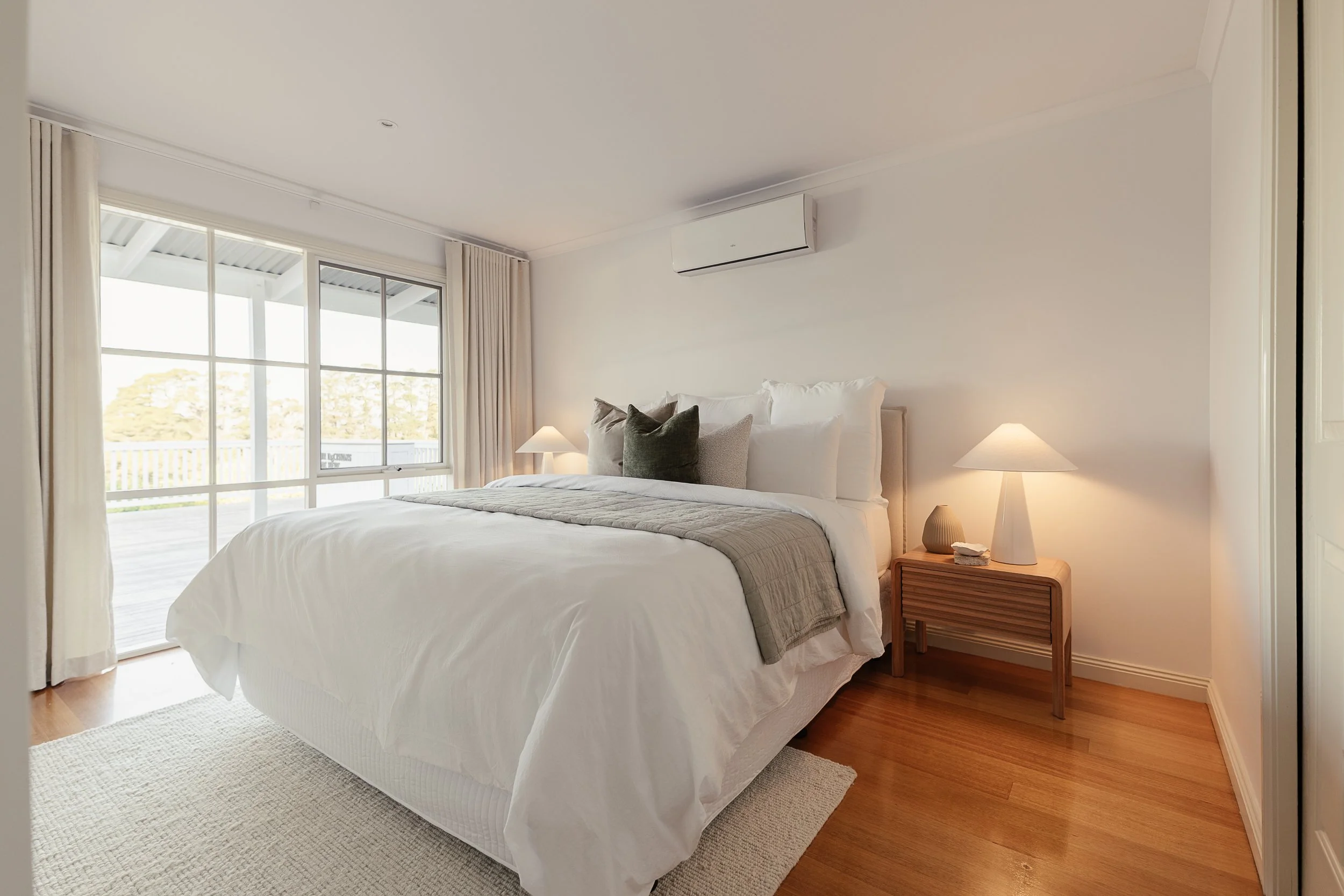 A neatly made bed with white bedding, multiple pillows including dark and light colors, a wooden nightstand with a beige lamp, and a window with beige curtains showing an outdoor balcony and trees.