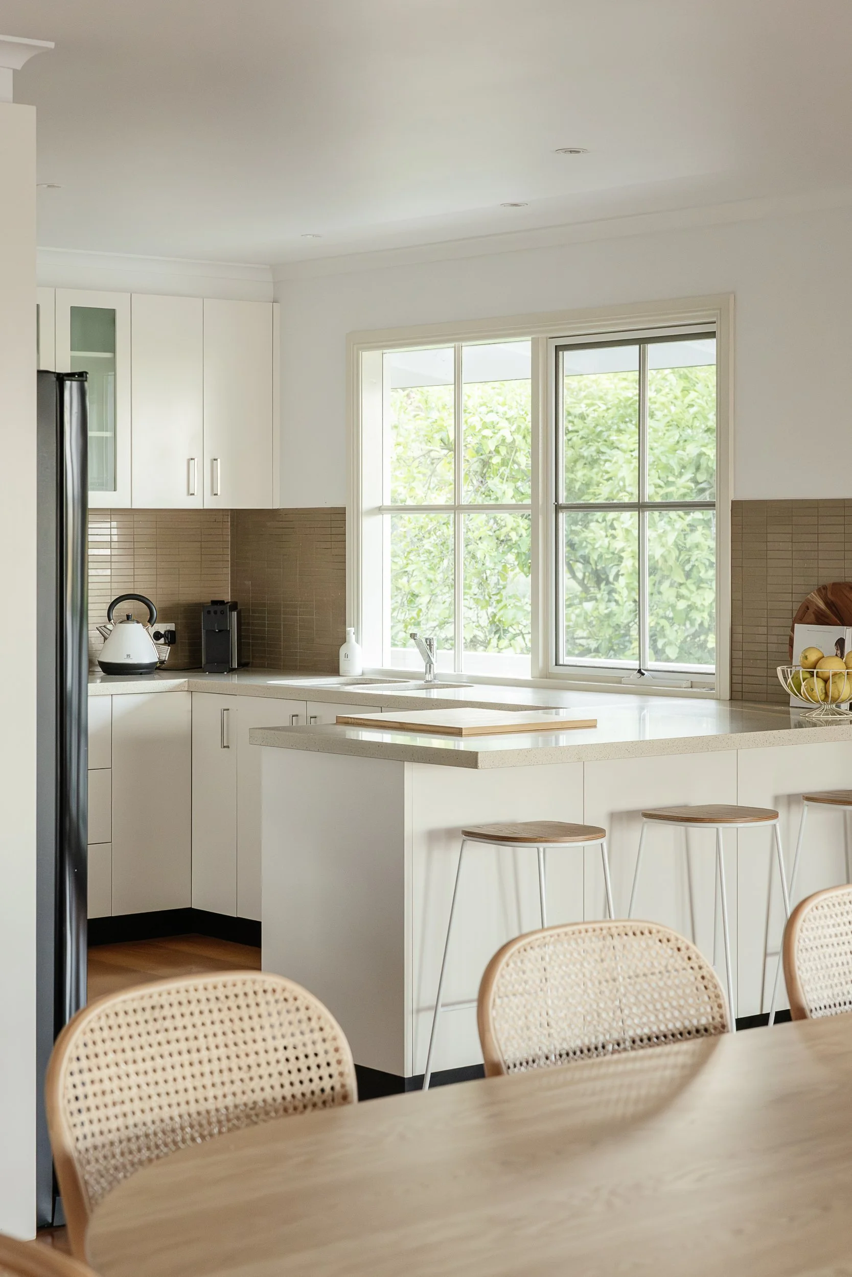 A modern kitchen with white cabinets, a large window with a view of green trees, a countertop with a white kettle and coffee machine, and wooden bar stools at a breakfast bar.