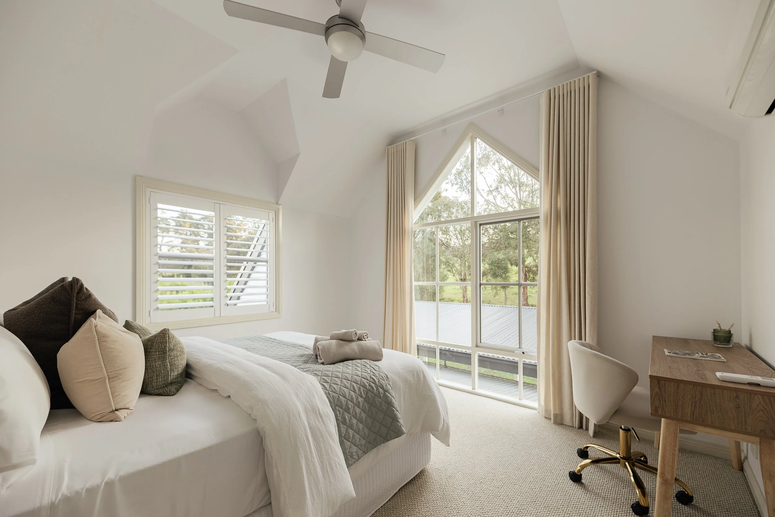 A bedroom with a large bed dressed in white linens and pillows, a window with white plantation shutters, tall beige curtains, a ceiling fan, a window view of trees, a wooden desk with a magazine and a green candle, a white chair, and a gold and black office chair.