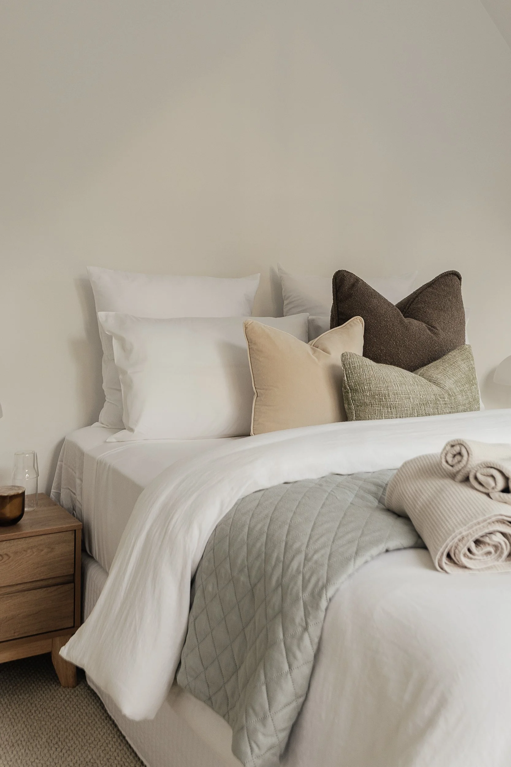 A neatly made bed with white bedding, multiple pillows including beige, dark brown, and light gray, a wooden nightstand with a glass and a bowl, and rolled towels on the bed, against a plain light-colored wall.