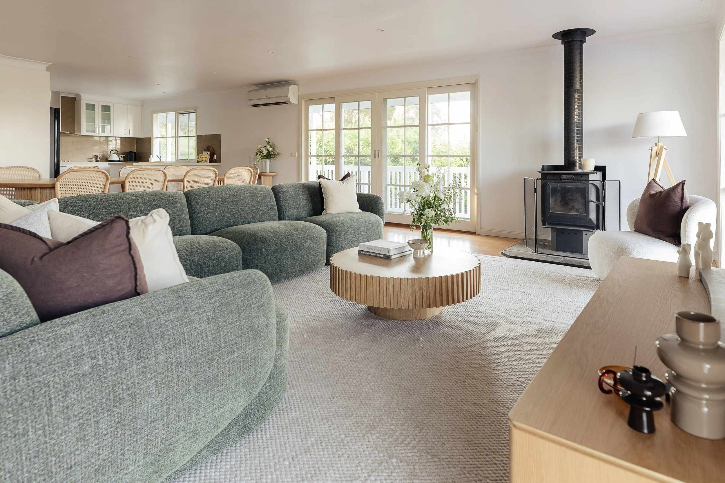 Living room with a green couch, a wooden coffee table with books and flowers, a wood stove, a white armchair, and large windows showing greenery outside.