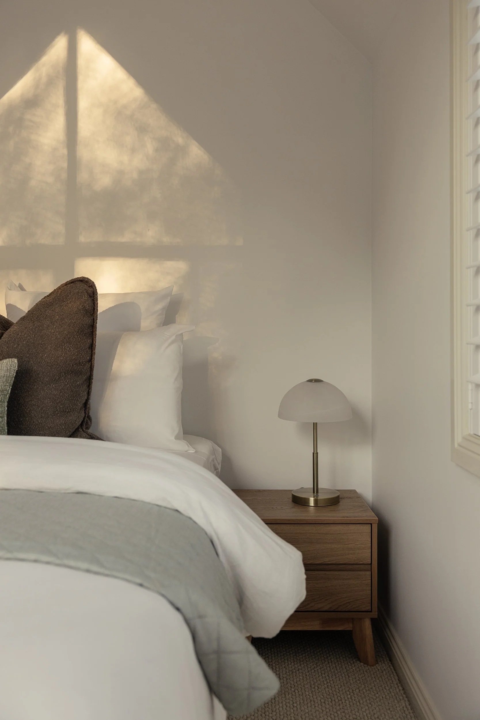 A bedroom with a bed, brown pillows, white and gray bedding, a wooden nightstand, and a white table lamp, with sunlight casting shadows through plantation shutters on the wall.