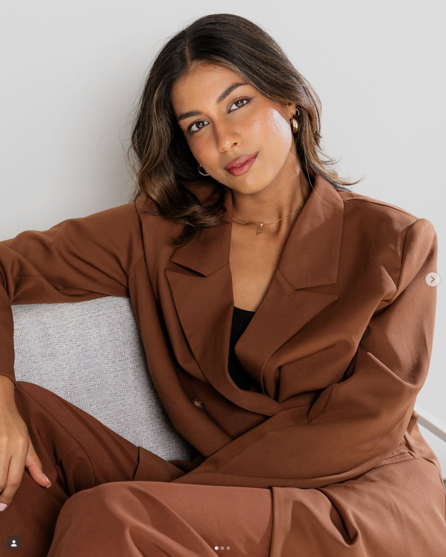 A woman with medium-length dark brown hair, brown eyes, and light brown skin wearing a brown jacket and gold jewelry sitting on a gray sofa.